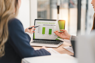 Two business professionals discussing carbon emissions reduction chart displayed on laptop screen...