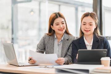 Two businesswomen in formal attire working together at desk with laptop, tablet, and documents in modern office setting, showcasing teamwork and collaboration