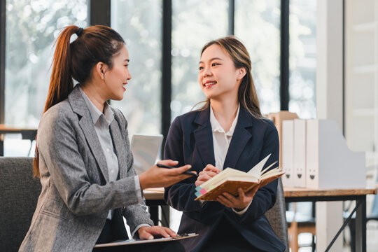 Two professional women in business attire are having friendly discussion in modern office setting, with one holding notebook and other gesturing while speaking