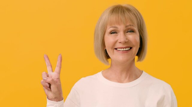 A cheerful woman with a short bob hairstyle flashes a bright smile while making a peace sign with her fingers. The vibrant yellow background enhances her joyful expression.