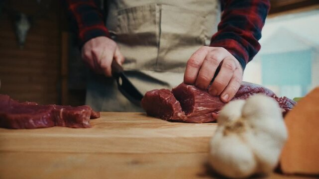 A person cutting fresh red meat on a wooden table, with garlic and bread in focus