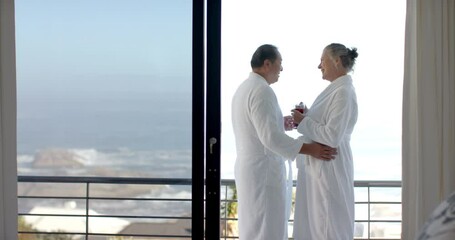 Senior couple in bathrobes enjoying ocean view and holding drinks on balcony, copy space