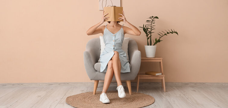 Pretty young woman covering face with book on armchair at home