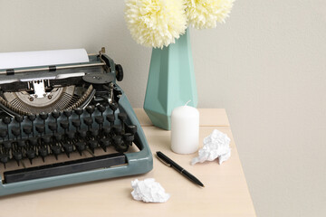 Vintage typewriter with crumpled paper, vase of flowers and candle on table near grey wall