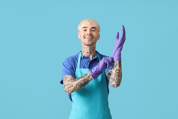 Young tattooed man putting rubber gloves on blue background