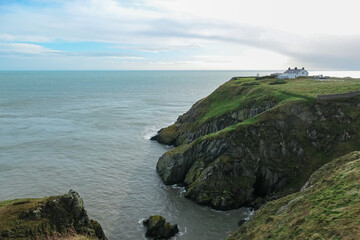 Dramatic coastal scene in Howth, Ireland. Steep cliffs plunging into grey choppy sea. Remote farm stands on distant headland. Overcast sky creating muted atmospheric light. Wild windswept beauty