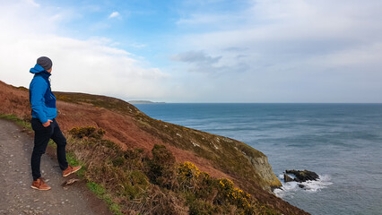 Man on scenic path winds along steep, grassy cliff overlooking grey sea under overcast sky. Coastline in peninsular village Howth in Dublin, Ireland. Peaceful solitude and coastal beauty, Irish coast