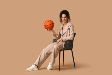 Female African-American basketball trainer in business suit with ball sitting on chair against...