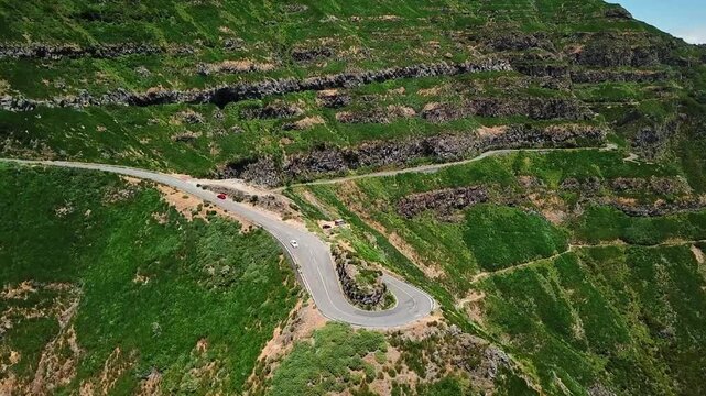 Aerial view of winding road at Miradouro Lombo do Mouro, Madeira, surrounded by lush green cliffs and dramatic terraced hillsides under clear blue sky, showing natural beauty and engineering marvels