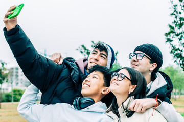 Four cheerful multi-ethnic students are taking a selfie together outdoors, embracing friendship and diversity, smiling and looking up at the smartphone