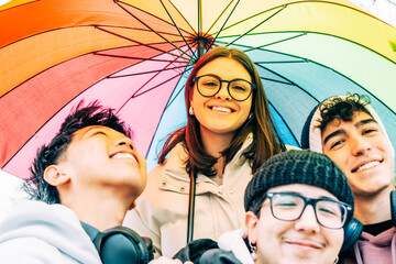Four cheerful multi-ethnic young students posing together under a vibrant rainbow umbrella, radiating joy, celebrating friendship, and embracing diversity in a lively outdoor setting