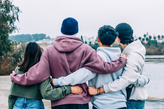 Four young multiracial friends are hugging each other while looking at the river, enjoying a moment of togetherness and friendship