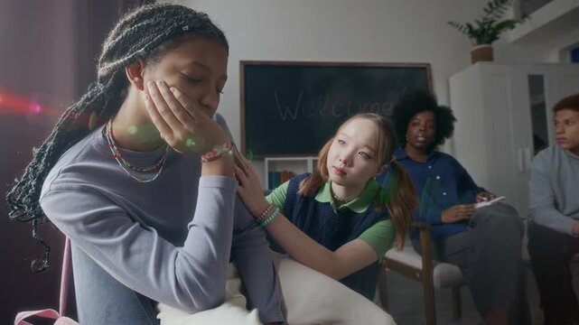 Medium shot of sad Black teen girl in despair surrounded by young Black female psychologist and multiethnic peers trying to support and create safe environment to talk about her crisis