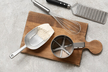 Wooden board with metal sieve mug, flour, whisk and grater on grunge background