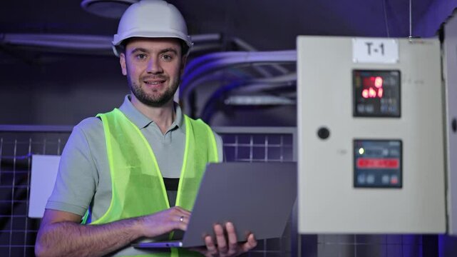 Electrical engineer uses laptop next to electrical switchboard. Professional man in hardhat and safety vest monitors equipment. Secure server room with digital control panel.