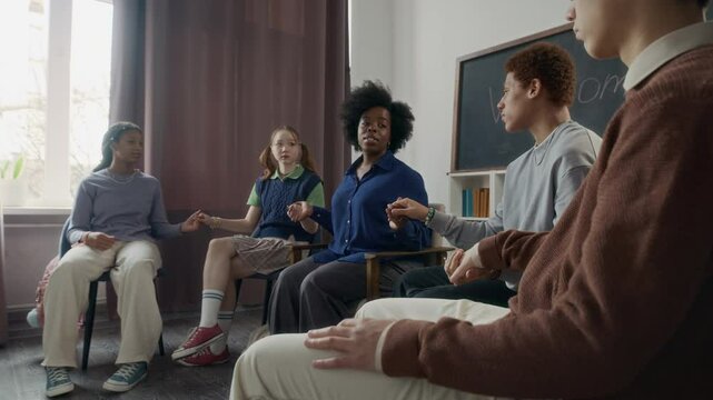 Medium wide shot of young African American psychologist leading group therapy session in modern office with multiethnic gen z kids and inviting them to hold hands together sitting in circle