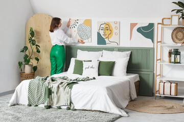 Young woman hanging painting on light wall in bedroom, back view
