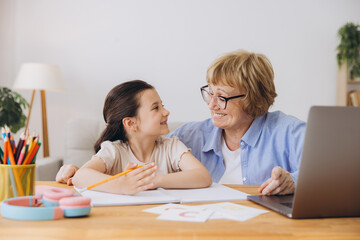 Happy grandma helping adorable preschool granddaughter school kid with homework task. Positive...