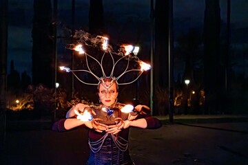 Woman fire performer juggling flaming torches while wearing elaborate headwear with flames at night