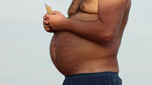 Adult man with big hairy stomach holds bag with salted nuts, his naked torso revealing effects of poor unbalanced diet. Excessive beer consumption and calorie surplus from junk food. Appetite control