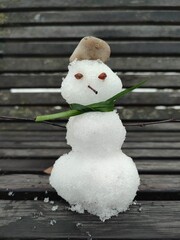 A small handmade snowman with a grass scarf, twig arms, and a pebble hat sitting on a wooden bench