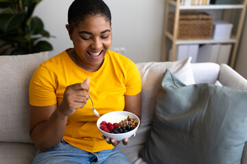 Happy black woman enjoying healthy breakfast with yogurt and berries on sofa