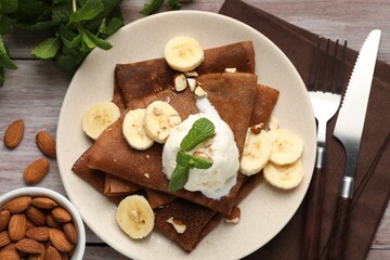 Delicious chocolate crepes with banana, nuts, mint and scoop of ice cream on wooden table, flat lay