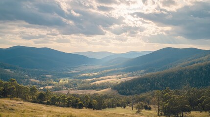 Obraz premium Sunlit mountain valley landscape with layered blue hills and sun rays breaking through clouds