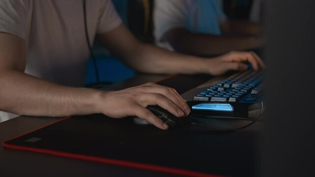 Gamer plays video game on PC at computer club with team, professional cyber sport tournament, E-sport championship. Focus on gaming mouse with RGB light keyboard on mat. Multiplayer shooter strategy