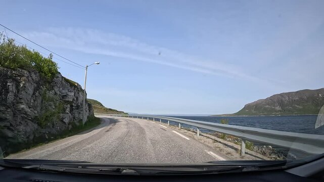 Driving through the tundra on E69 in northern Norway. Rocky hills with sparse trees and grass rise on the left, while the sea stretches along the right, showcasing raw and rugged nature.
