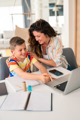 Caucasian mother in casual attire assists her elementary-aged son with schoolwork at home, using...