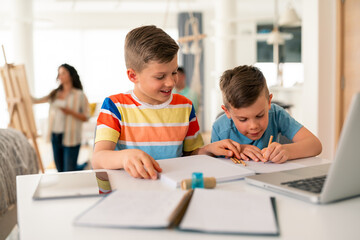 Fototapeta premium Two Caucasian brothers in casual attire collaborate on schoolwork using laptop and books at bright home study space, demonstrating modern learning methods.