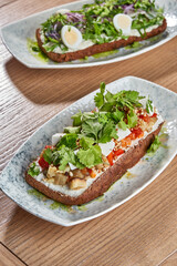 A vibrant and colorful spread of two smorrebrod, traditional Danish open-faced sandwiches, topped with fresh vegetables, herbs, and creamy spreads.