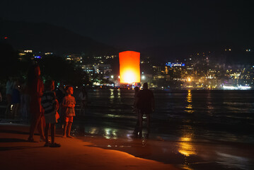A glowing red lantern floats above a beach in Phuket, Thailand, as people gather. Town lights...