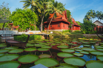 Thai style red wooden buildings with sloped roofs surrounded by greenery, a pond with lily pads, a...