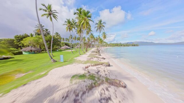 FPV Flight Through Coconut Trees at Playa del Aserradero, Saman&aacute;, Highlighting the Beach, Green Grass, and Gazebos on a Sunny Day.