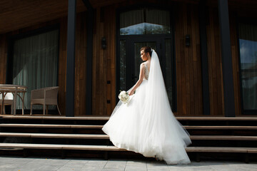 the bride is turned sideways, holding a bouquet of white flowers. The dress is long, with a full...