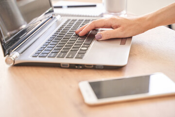 Caucasian female adult using laptop with smartphone on wooden desk in bright workspace.