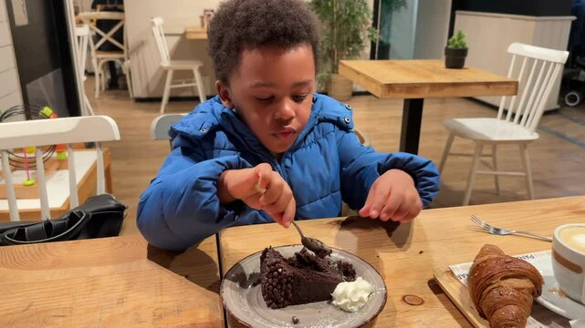 lovely 4-year-old biracial black kid eating a chocolate cake by himself with a spoon inside a cefetria seated in a wooden table. We is wearing curly hair and winter clothing. 4K