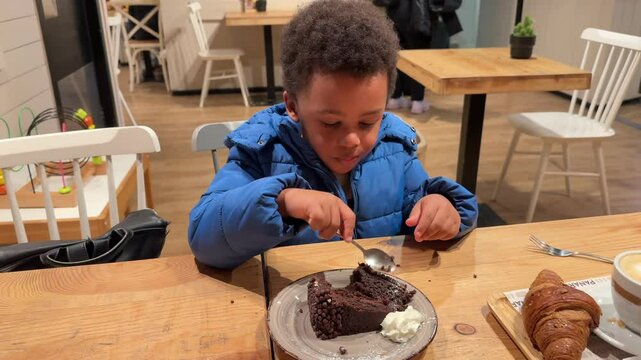 4-year-old glutton biracial black kid eating a chocolate cake by himself seated in a wooden table inside a cafeteria. He is wearing curly hair and winter clothing. 4K