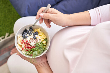 Pregnant asian female enjoying healthy fruit and yogurt bowl in cozy setting.