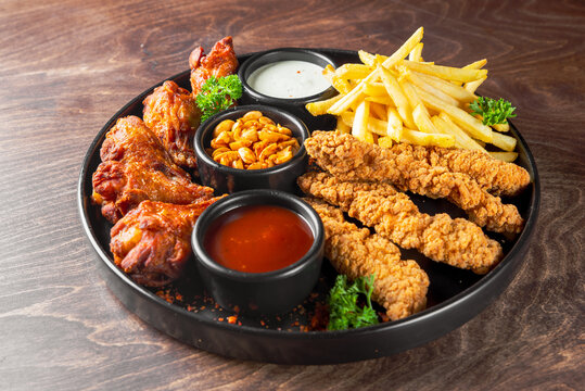 A delicious platter featuring crispy fried chicken tenders, spicy chicken wings, golden French fries, and peanuts, accompanied by dipping sauces, garnished with fresh parsley on a wooden table.