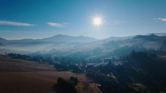 Aerial view of sunlight streaming over a misty valley, rolling hills, and scattered trees under a crisp, clear blue sky. (4K)