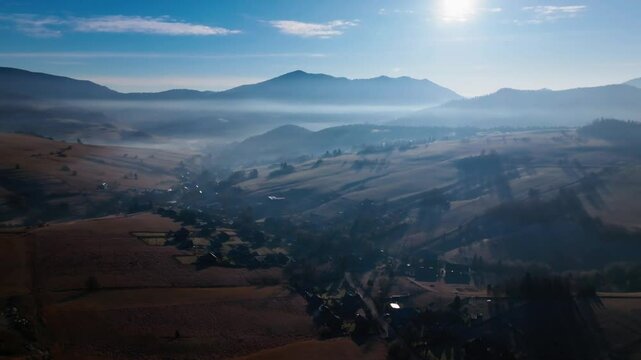 Stunning aerial view of mist rolling through a sunlit valley, surrounded by picturesque hills and scattered rural homes under a bright blue winter sky. (4K)