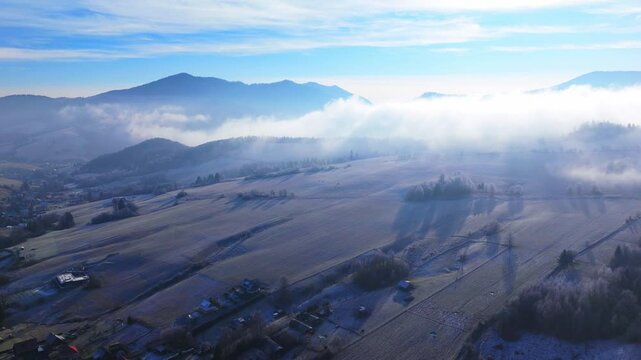 Aerial hyperlapse video of rolling hills, fog drifting over frosty fields, and scattered rural villages under a bright blue sky.