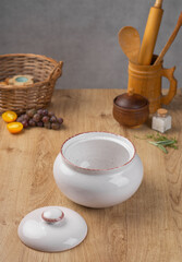 White ceramic clay pot with lid for roasting meat and vegetables stands on kitchen wooden table