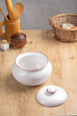 White ceramic clay pot with lid for roasting meat and vegetables stands on kitchen wooden table