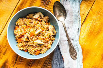 Boiled spelled or spelled porridge with mushrooms, carrots and spinach in a blue bowl on a wooden background with a napkin and spoon. Healthy eating concept. Vegetarian dish. Copyspace