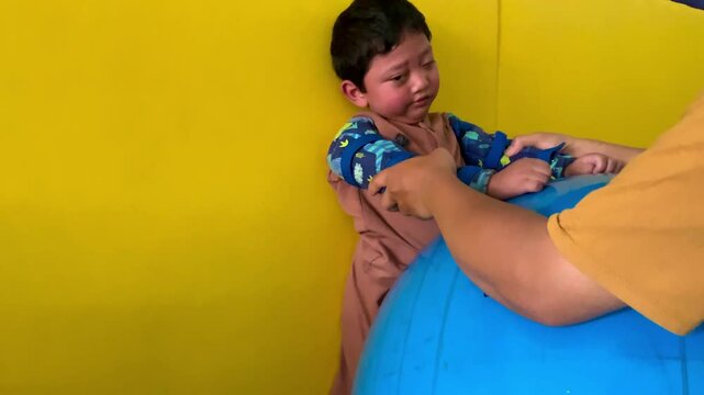 The boy who lives with cerebral palsy is learning to stand up to the wall covered in the mattress and flanked by a soft blue ball assisted by a professional physiotherapist