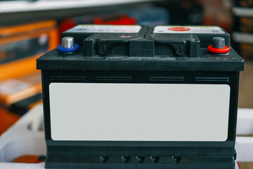 Black car battery with blank label on the front, standing on a white surface in a store. Blurred background with shelves full of products. Perfect for branding and automotive-themed designs